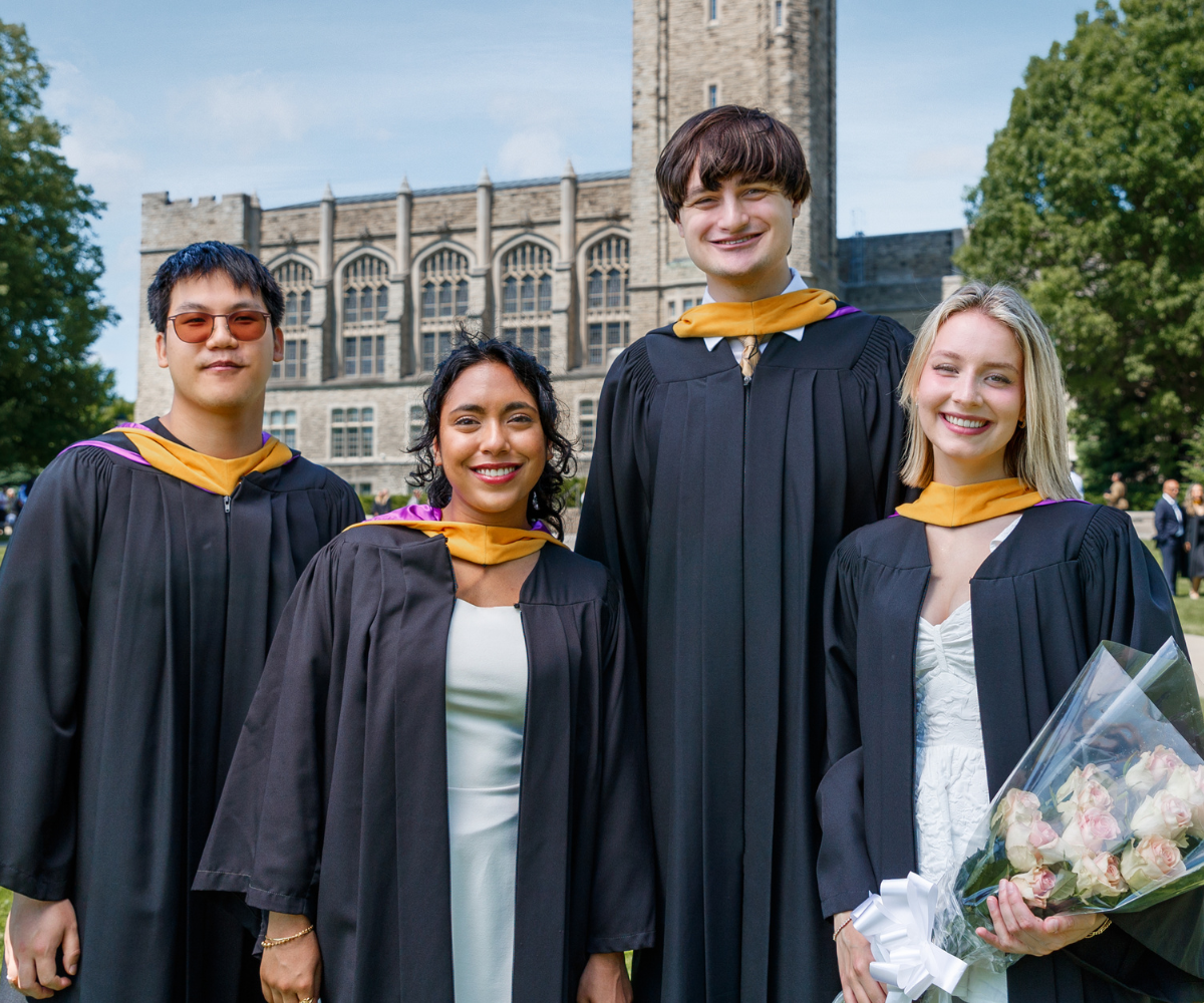 A group of graduates wearing academic gowns and hoods pose together on a grassy area in front of a large stone campus building. One person holds a bouquet of flowers.