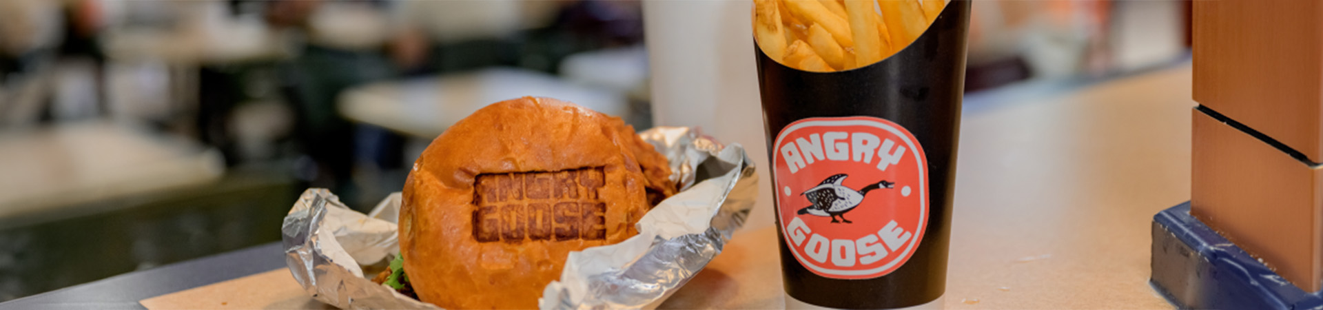 Burger wrapped in foil beside fries and a drink with an Angry Goose logo, on a table in a busy food court.
