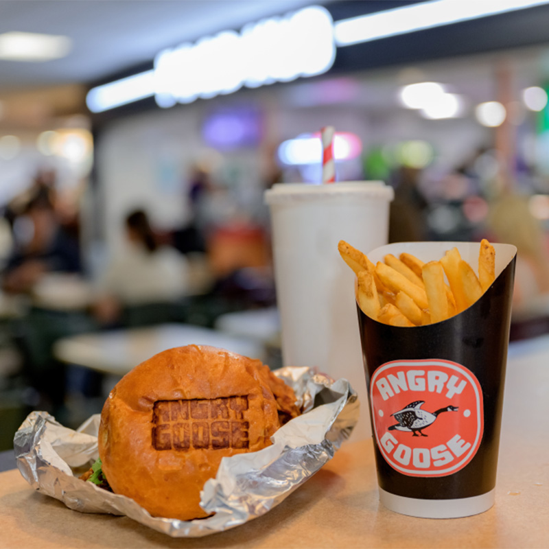 Burger wrapped in foil beside fries and a drink with an Angry Goose logo, on a table in a busy food court.