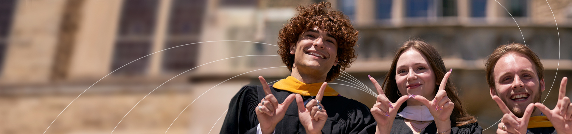 Three graduates in academic gowns standing outdoors in front of a stone campus building, smiling and holding up hand signs with raised index fingers under bright daylight.