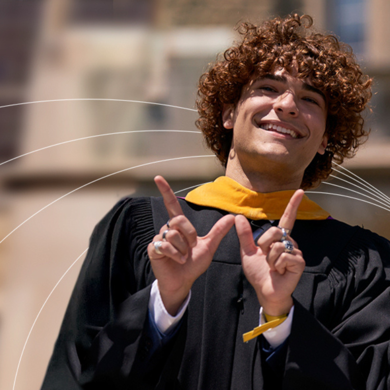 Three graduates in academic gowns standing outdoors in front of a stone campus building, smiling and holding up hand signs with raised index fingers under bright daylight.