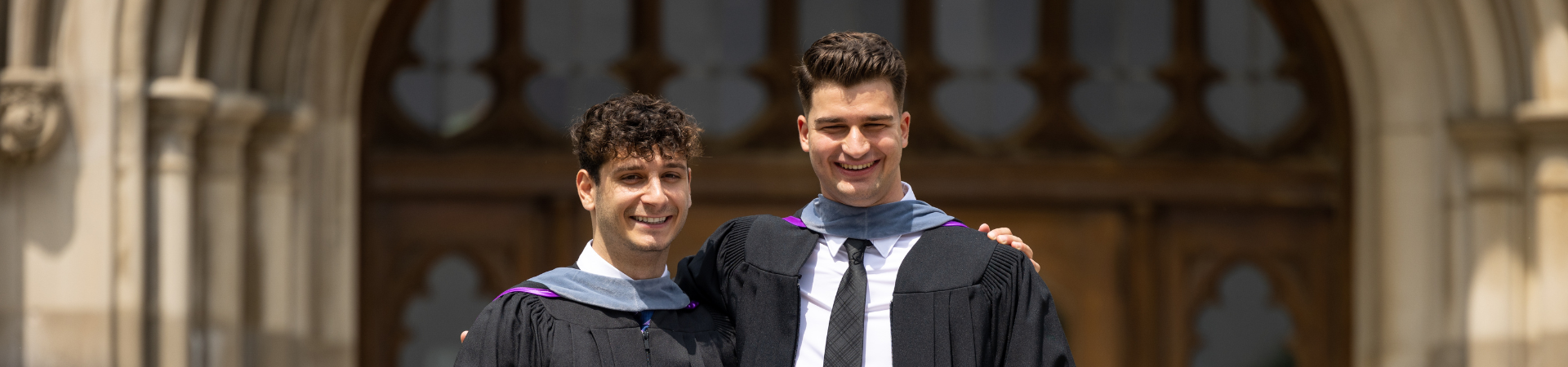 Two male graduates posing for a photo together side by side with their arms around one another's shoulders
