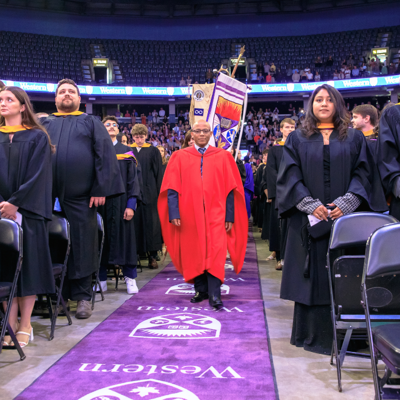 Two graduates seated at their ceremony wearing academic regalia and smiling at a camera