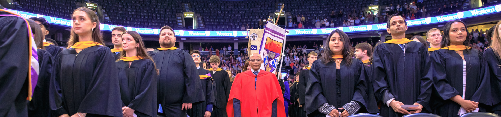 Two graduates seated at their ceremony wearing academic regalia and smiling at a camera