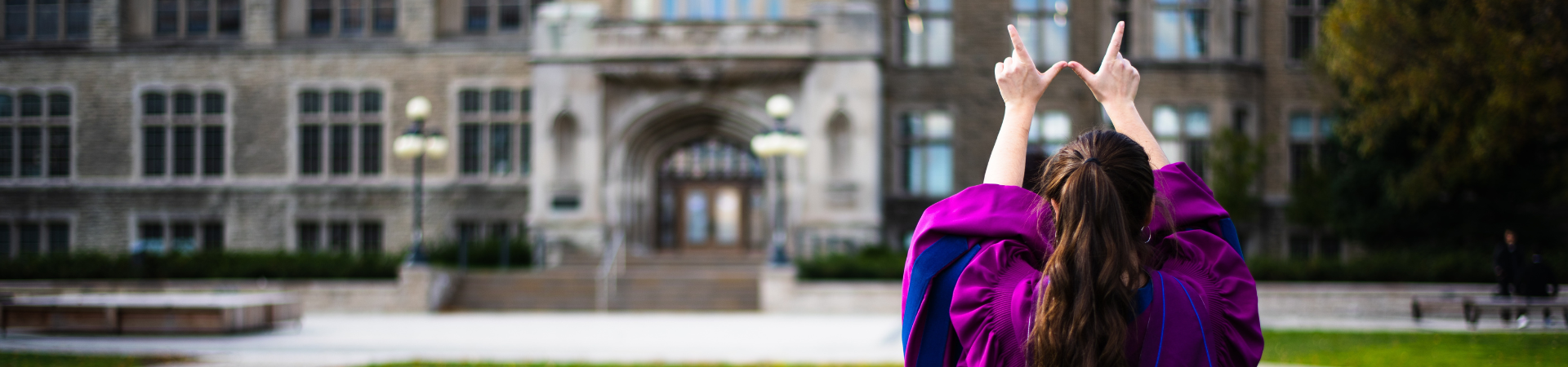 A graduate posing infront of University College making a "w" with their hands