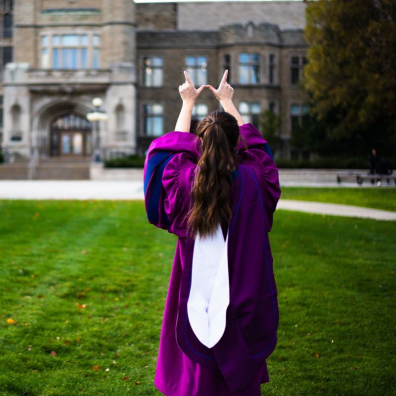 A graduate posing infront of University College making a "w" with their hands