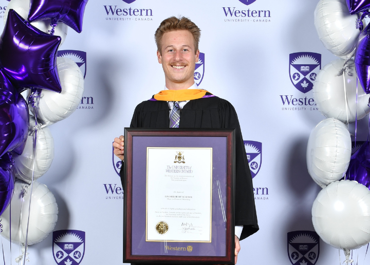 A graduate standing in front of a Western backdrop holding their framed diploma