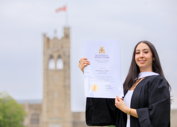 A female graduate standing outside in front of University College on campus posing with their degree