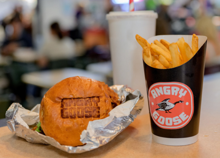 Burger wrapped in foil beside fries and a drink with an Angry Goose logo, on a table in a busy food court.