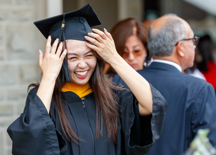 A graduate standing outside putting on a graduation cap and smiling
